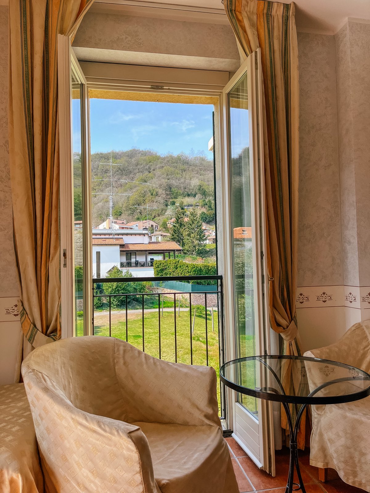 Balcony Room: an armchair and a glass table at the open balcony doors, the garden and hills outside.