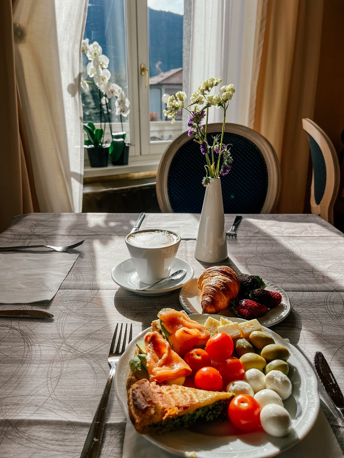 A laid breakfast table by a tall window: cappuccino, croissant, a plate of fruit and pastries.